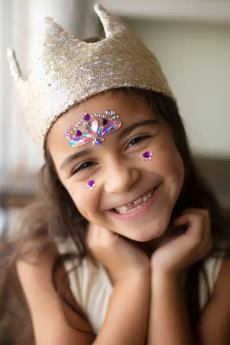 Een meisje draagt roze gezicht stickers met kristallen prinsespatronen op haar gezicht en een glinsterende kroon.