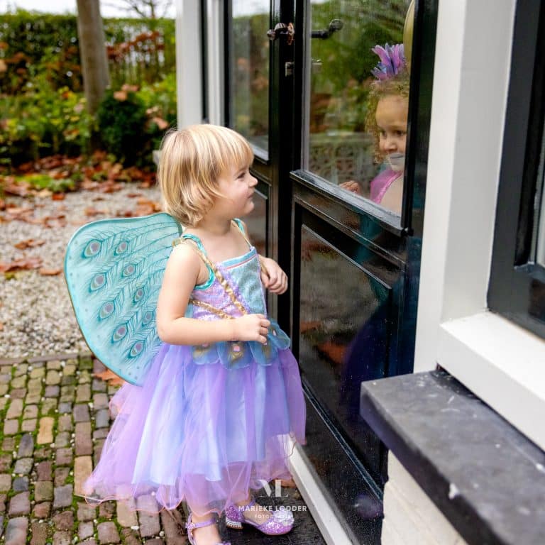 Bij de voordeur staat een meisje in de paarse elfen jurk Tamara met turquoise pauwenvleugels en paarse tule rok