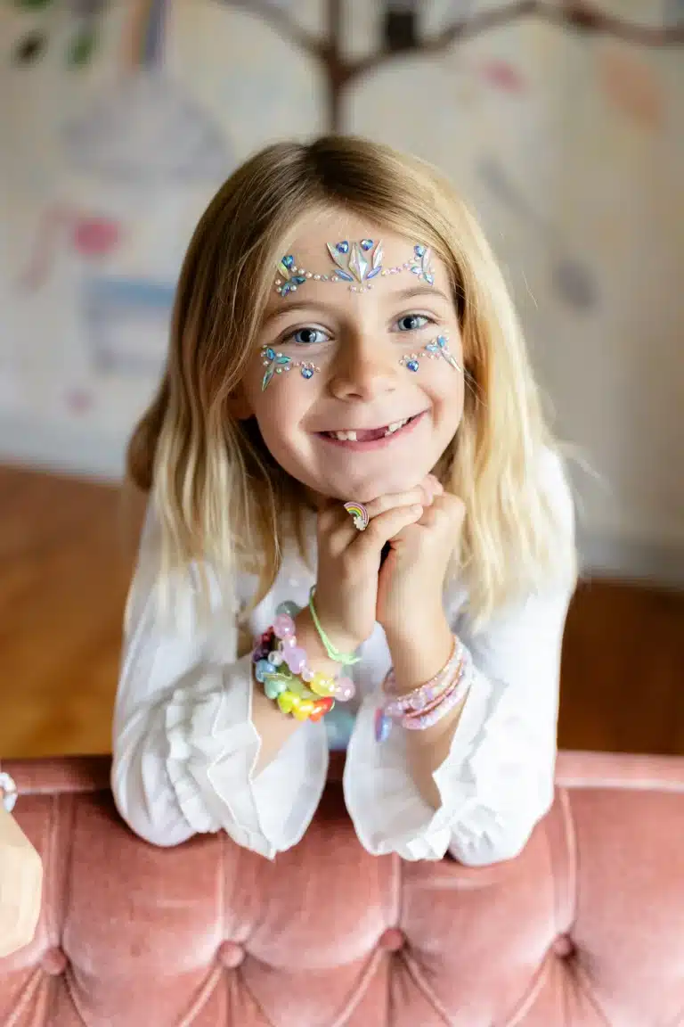 Lachend blond meisje zit binnen met haar handen onder de kin en draagt gezichtstickers kristallen ijsprinses