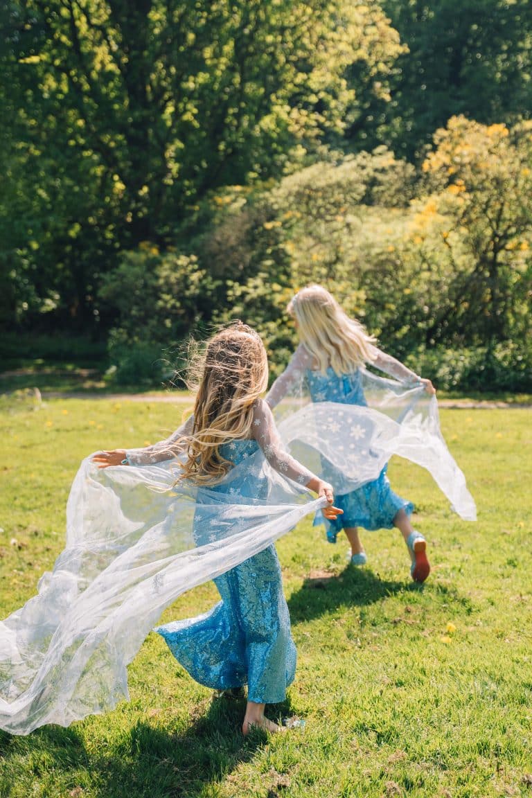Twee meisjes dragen blauwe Elsa jurken met glinsterende rok en transparante sleep terwijl ze over het gras rennen