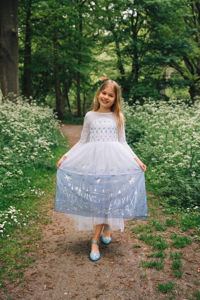 Meisje draagt een wit frozen jurkje elsa met glinsterende rok en blauwe prinsessenschoenen op een bospad