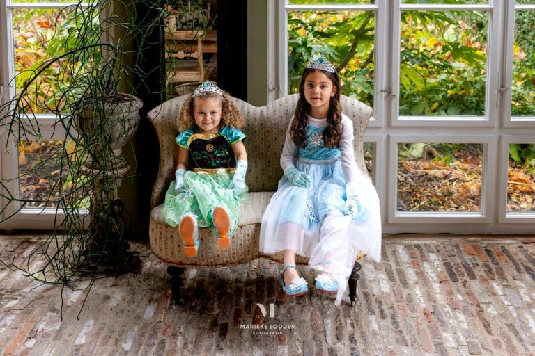 Twee meisjes dragen de elsa en anna jurk uit de combideal, zittend in een fauteuil met kroontjes en handschoenen.