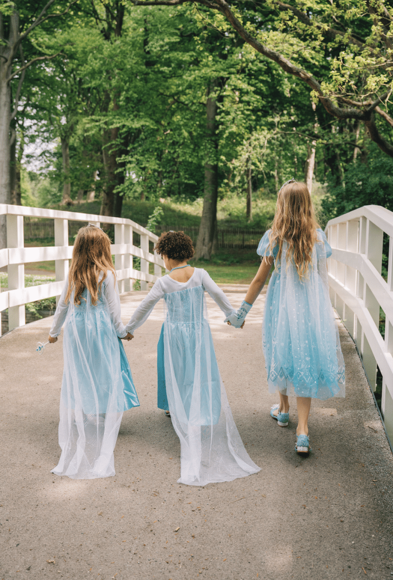 Drie meisjes dragen een blauwe Elsa jurk en lopen hand in hand over een witte brug in het bos