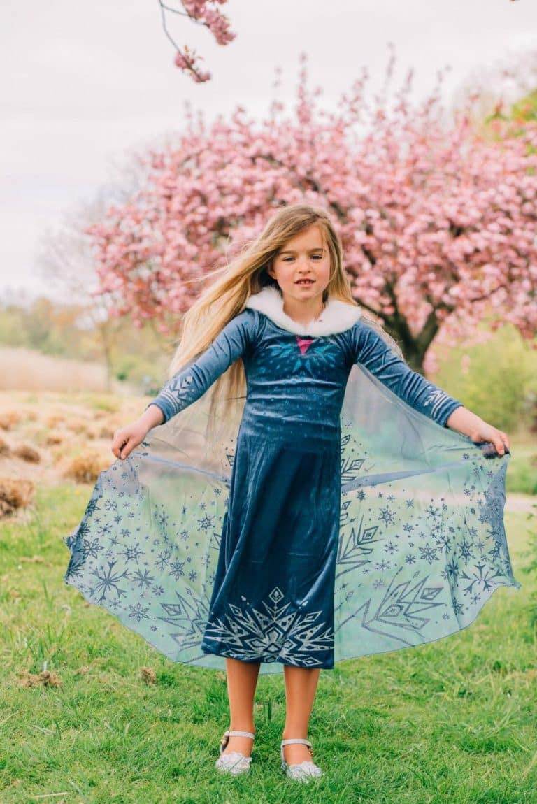 Meisje draagt een blauw Frozen kleedje met witte kraag en doorschijnende sneeuwvlokcape in een bloeiende tuin