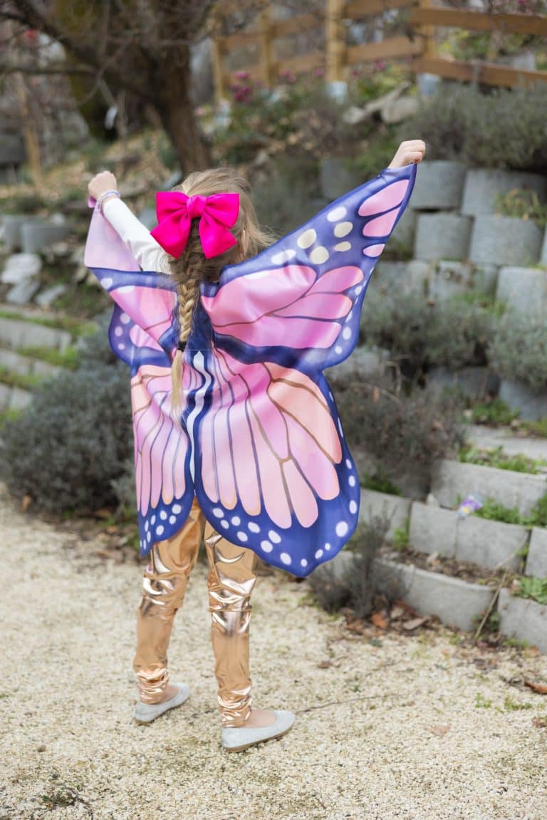 Meisje met de butterfly wings set: roze en paarse vlindervleugels met blauwe randen, van achteren gezien in de tuin.