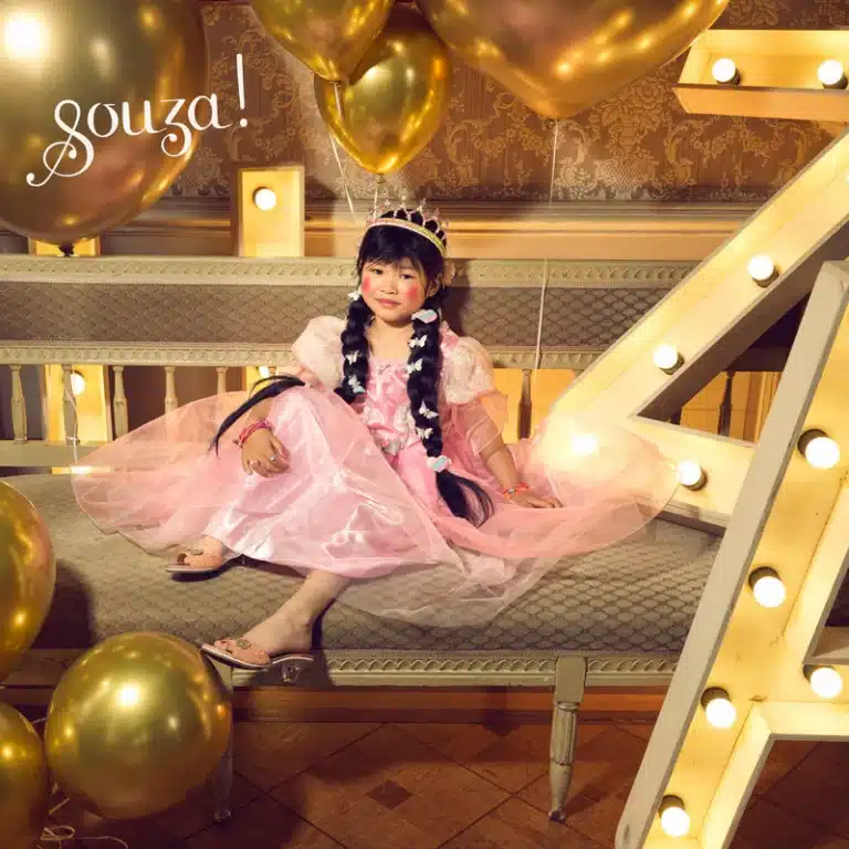 Meisje draagt de roze Souza jurk Annerose met wijde tule rok en pofmouwen, zittend tussen gouden ballonnen.