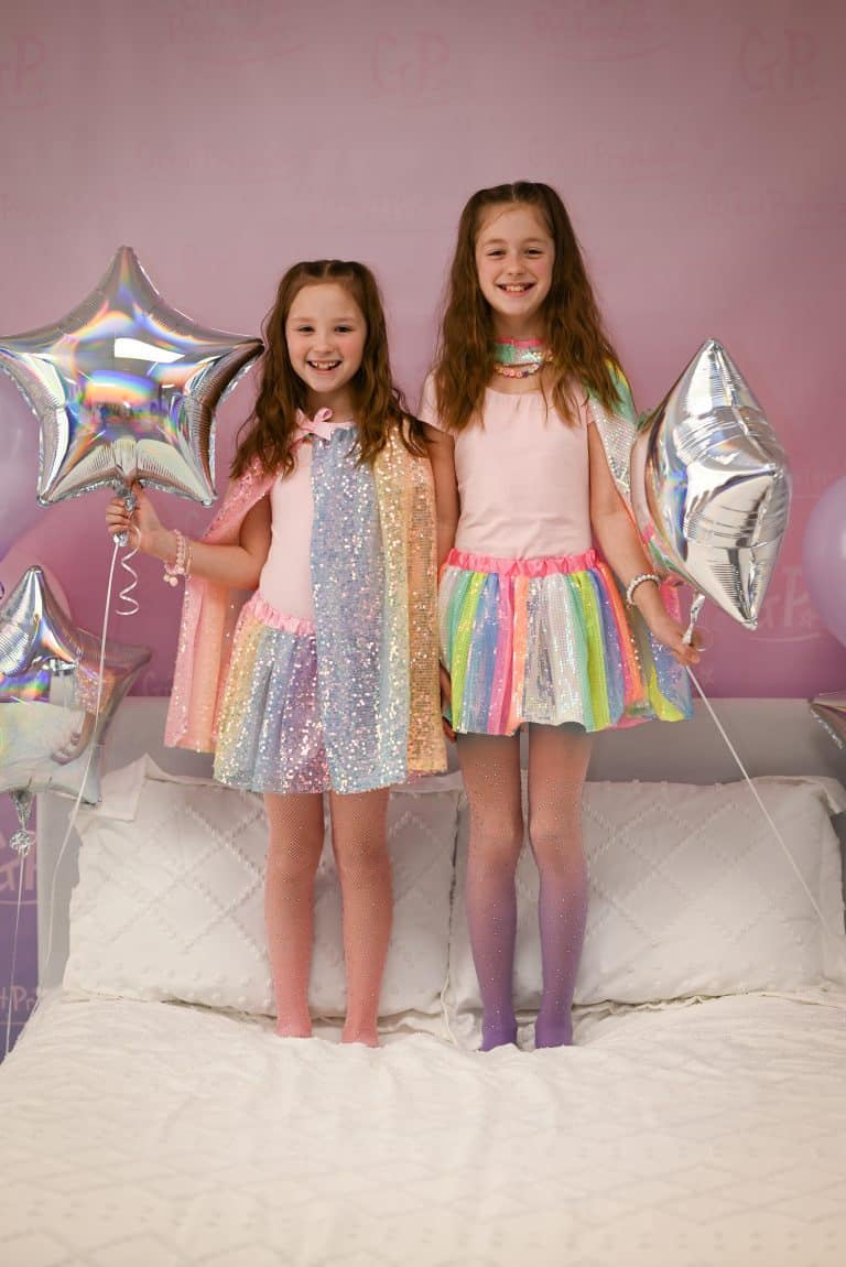 Een meisje draagt het Great Pretenders regenboog rokje met glanzende gekleurde banen, naast een meisje in een paillettenjurk.