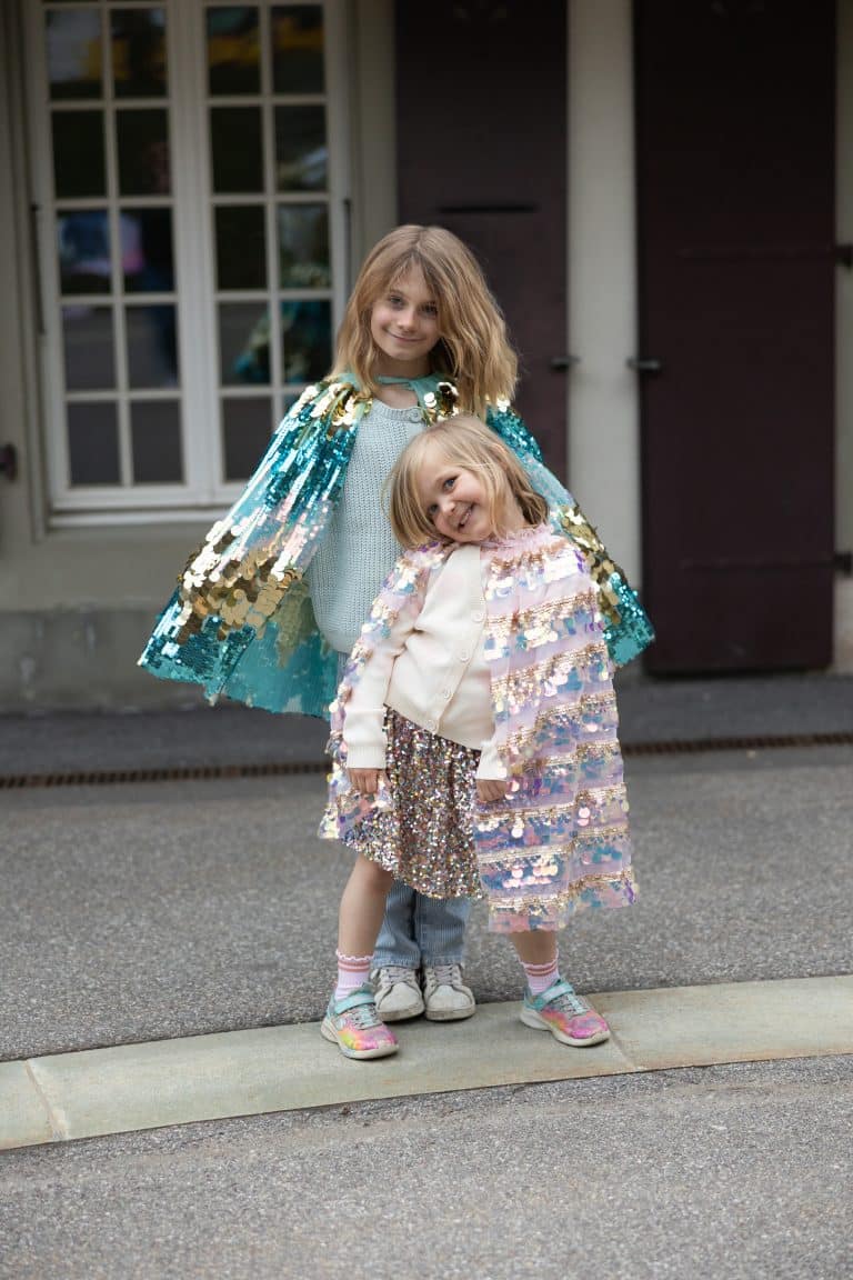 Twee meisjes dragen Great Pretenders capes; de voorste draagt een roze pailletten cape buiten