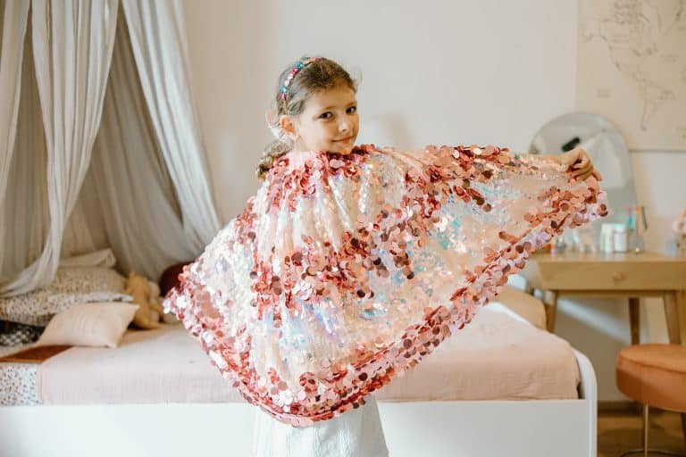Meisje draagt een lichtroze Great Pretenders cape met glanzende pailletten en houdt de stof aan beide kanten uitgespreid