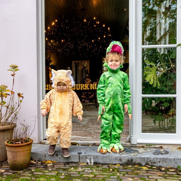 Kind draagt een groen dino carnavalspak met open bek en klauwen naast een kind in een tijgerpak bij de voordeur.