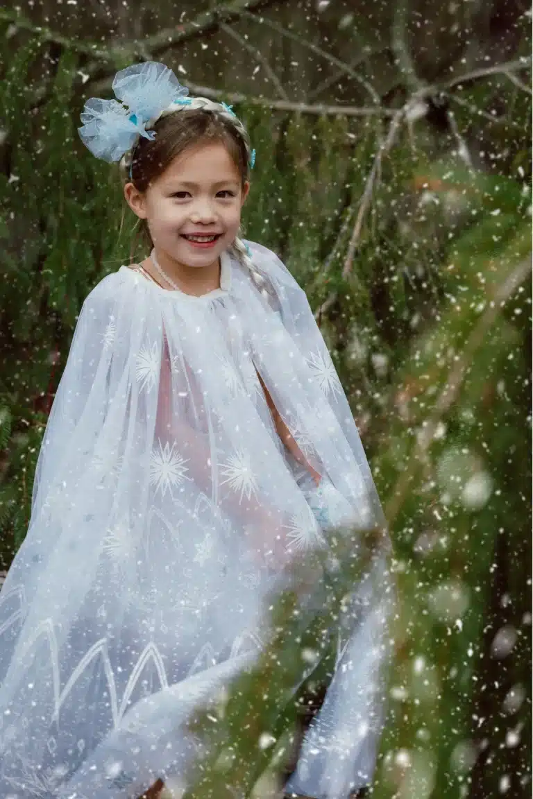 Van dichtbij toont een meisje de lichtblauwe Great Pretenders Elsa cape met sneeuwvlokken, terwijl ze die spreidt in het bos.