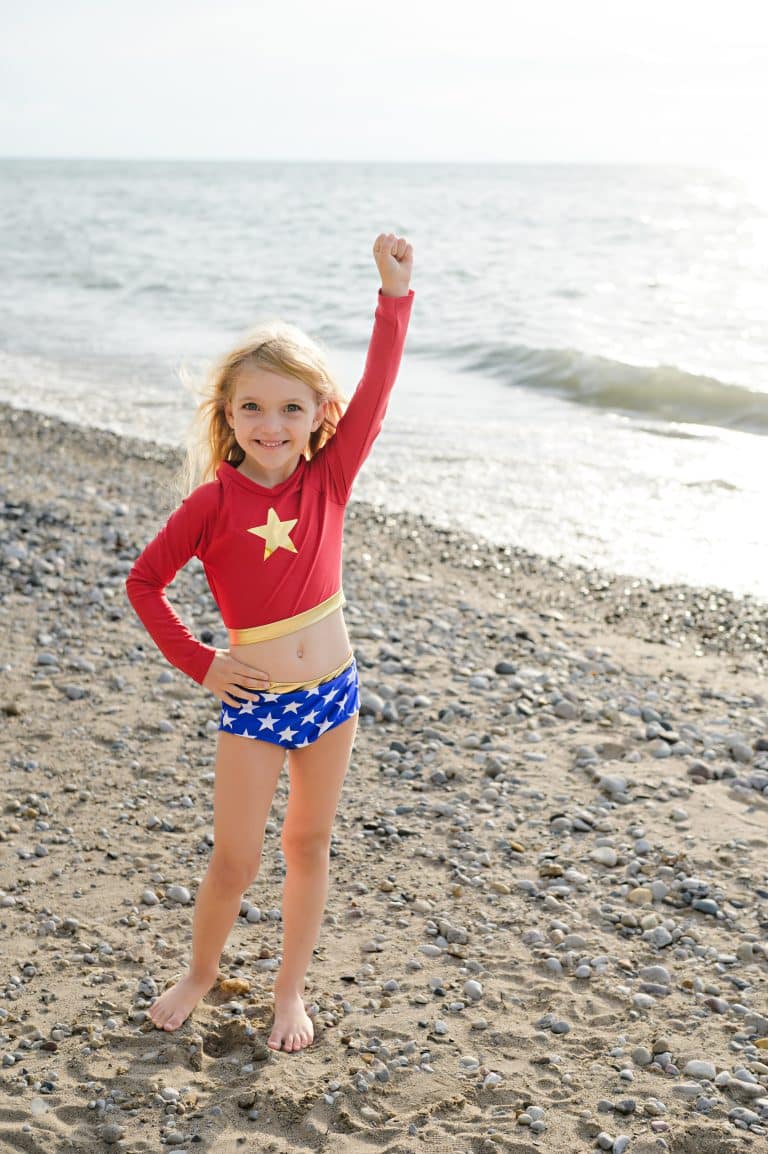 Met een opgeheven vuist poseert een meisje in het Super Girl badpakje met rode top en ster op het kiezelstrand.