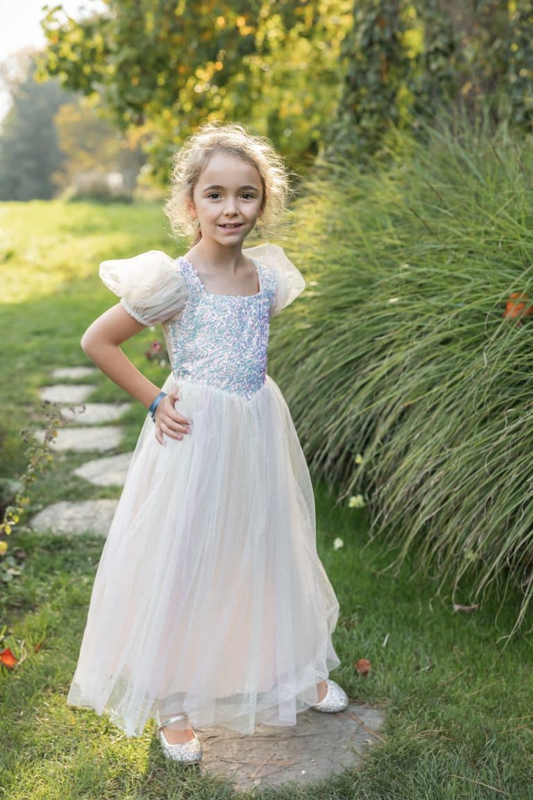 Op een tuinpad poseert een meisje in de witte Sparkle prinsessenjurk, met de handen in haar zij en glinsterende schoenen.