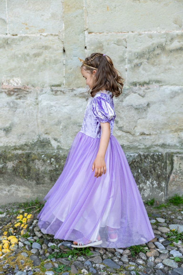 Meisje draagt een paarse Rapunzel verkleedjurk met lange tule rok en pofmouwen, buiten bij een stenen muur.