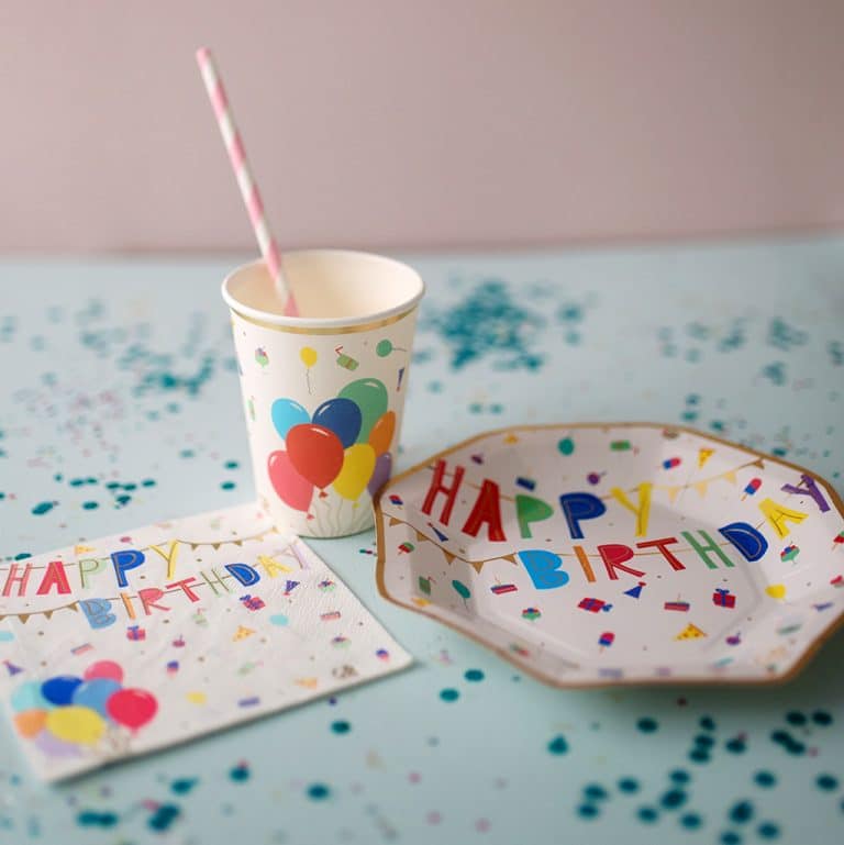 Happy Birthday beker van Great Pretenders met kleurrijke ballonnen, naast een bord en servet met verjaardagstekst