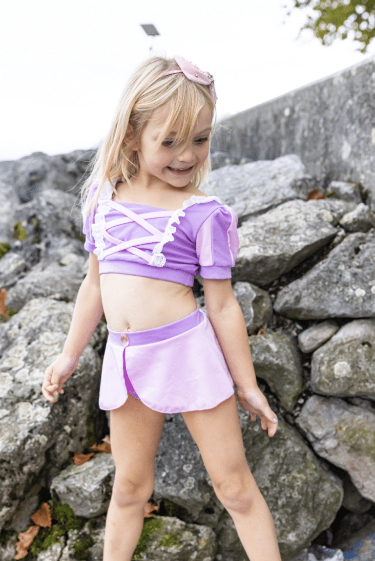 Meisje draagt een paarse rapunzel bikini met wit afgezet topje en rokje tussen rotsen