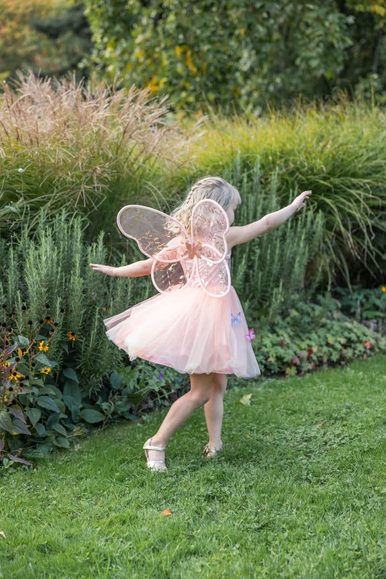 Meisje draagt roze fee vleugels met transparante glitterstof in een bloeiende tuin, gezien van achteren