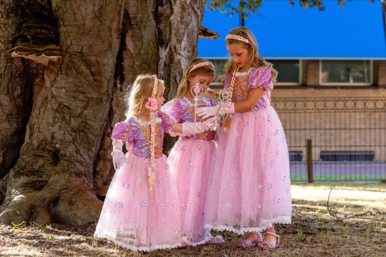 Drie meisjes dragen roze Rapunzel prinsessenjurken met bloemenprint, glitterdetails en lange tule rokken onder een boom