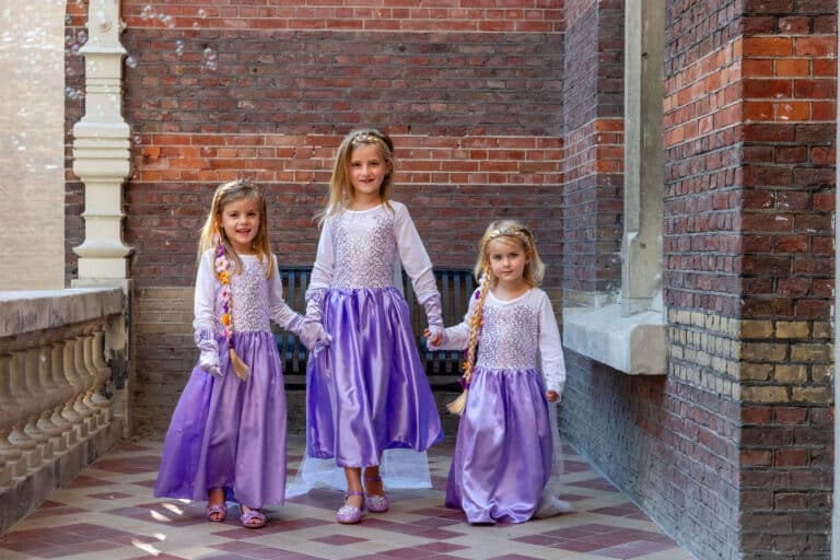 Drie meisjes dragen paarse Elsa jurken met glanzende lange rokken en witte mouwen in een bakstenen galerij.